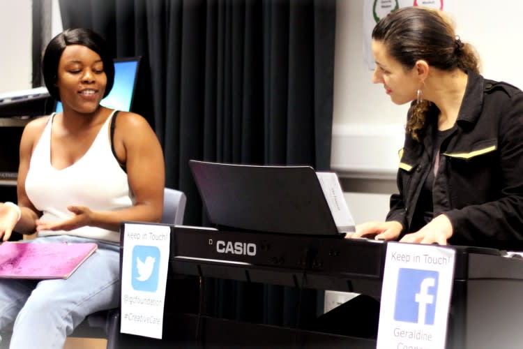 Two women sat at a piano, one is playing the other watching