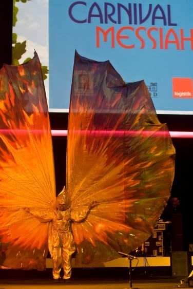 A performer stands onstage, arms raised presenting huge golden, yellow wings for Carnival Messiah
